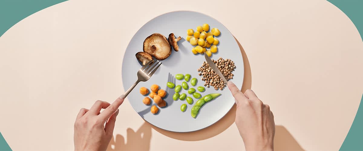 Eine Person hält eine Gabel und ein Messer über einem Teller mit verschiedenen Gemüsesorten, darunter Pilze, Süßlupine, Kichererbsen, Edamame und Nutzhanf