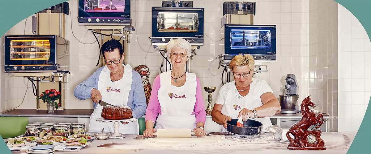 Drei Omas stehen in einer Küche und tragen weiße Schürzen mit dem Schriftzug „Elisabeth“. Eine Oma links schneidet einen Kuchen, die mittlere Oma hält ein Nudelholz, und die rechte Oma füllt eine Backform.