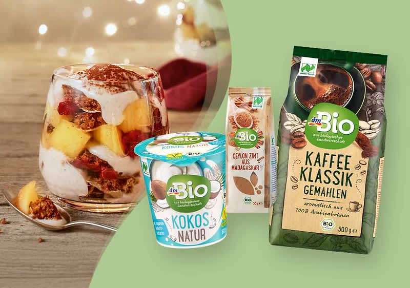 Links ein Dessertglas mit Schichten aus Joghurt, Müsli, Ananasstücken und Himbeeren, rechts drei dmBio-Produkte: ein Kokosjoghurt, eine Packung Ceylon-Zimt und eine Packung gemahlener Kaffee