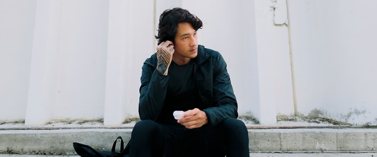 Ein Mann in dunkler Kleidung mit mittelangen schwarzen Haaren sitzt auf einer Treppe und hält ein weißes Case für Kopfhörer in der Hand, während er ein Kopfhörer in ein Ohr einsetzt.