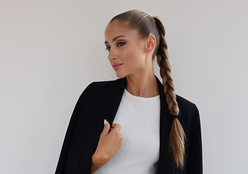 Eine Person mit einem langen, geflochtenen Zopf trägt ein schwarzes Jackett über einem weißen Oberteil und hat auffällig geschminkte Siren Eyes.