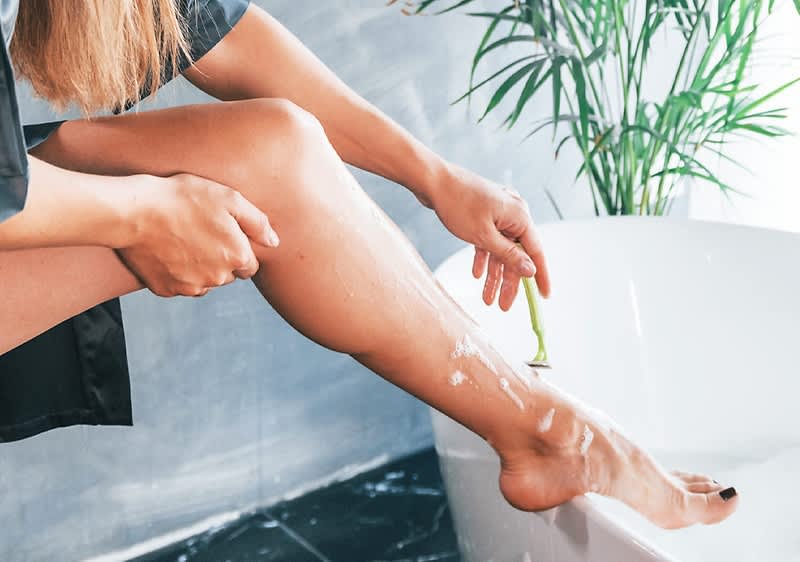 Eine Person rasiert ihr Bein mit einem grünen Rasierer, während sie am Rand einer Badewanne sitzt. Im Hintergrund steht eine grüne Pflanze.