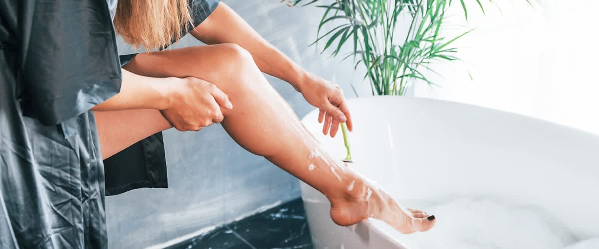 Eine Person rasiert ihr Bein mit einem grünen Rasierer, während sie am Rand einer Badewanne sitzt. Im Hintergrund steht eine grüne Pflanze.