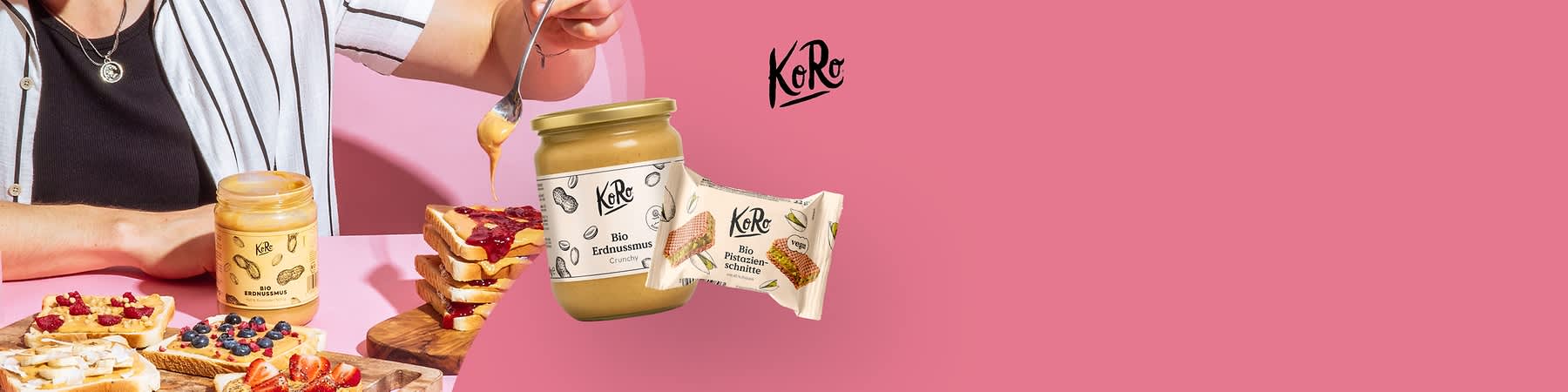 Ein Glas mit der Aufschrift „Bio Erdnussmus Crunchy“ neben einer Verpackung mit der Aufschrift „Bio Pistazienschnitte“ und dem KoRo-Logo auf rosa Hintergrund