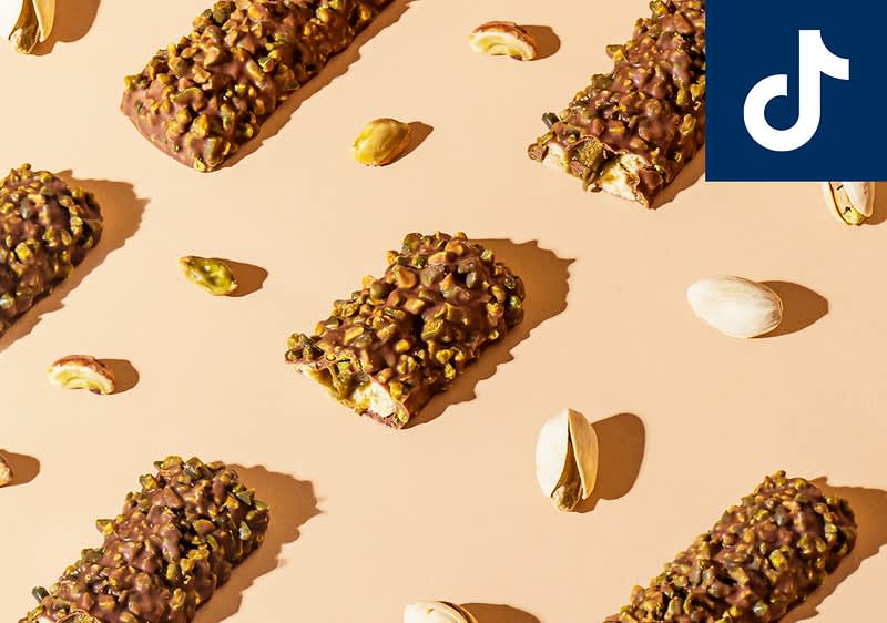 Mehrere Schokoriegel mit Pistazienstücken liegen auf einer beigen Fläche, umgeben von ganzen und geöffneten Pistazien. Oben rechts ist das Logo von TikTok.