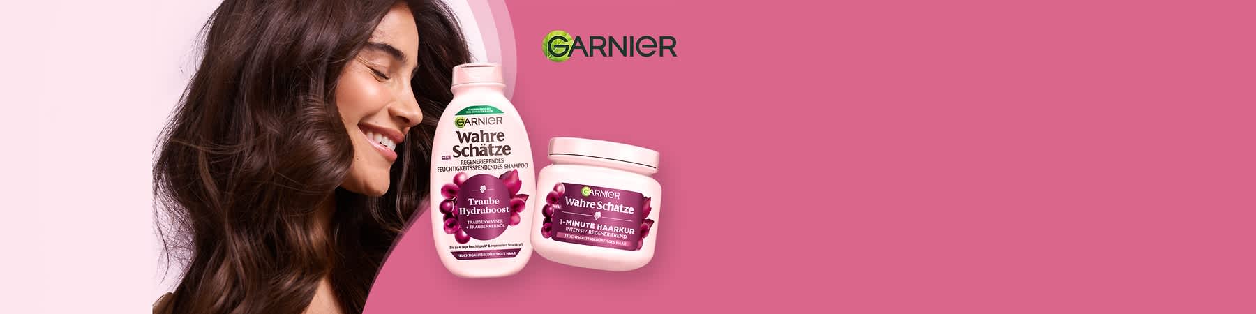 Zwei Produktverpackungen von Garnier Wahre Schätze auf rosa Hintergrund, links eine hell-rosa Flasche mit der Aufschrift „Traube Hydrabost regenerierendes feuchtigkeitsspendendes Shampoo“, rechts ein hell-rosa Tiegel mit der Aufschrift „1-Minute Haarkur intensiv regenerierend“. Links im Bild eine lächelnde Frau mit gesundem, braunem Haar