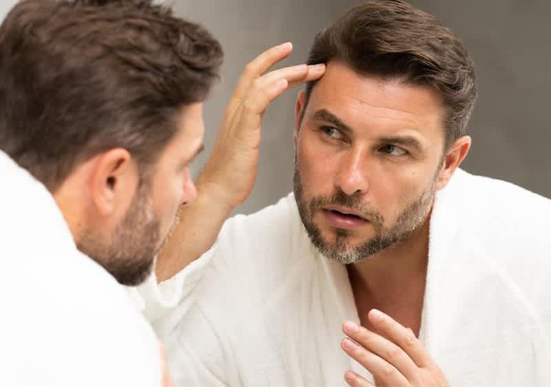 Ein Mann in einem weißen Bademantel betrachtet sich im Spiegel und frisiert seine Haare, um Geheimratsecken zu kaschieren