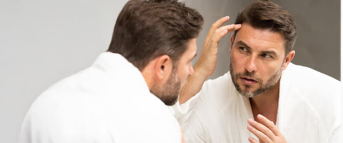 Ein Mann in einem weißen Bademantel betrachtet sich im Spiegel und frisiert seine Haare, um Geheimratsecken zu kaschieren