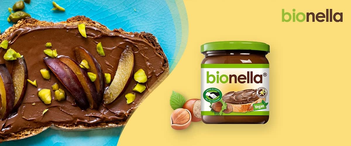 Ein Glas der Marke bionella mit grünem Deckel und der Aufschrift „Vegan“ neben einem Brot mit Schokoladencreme und Pistazienstücken