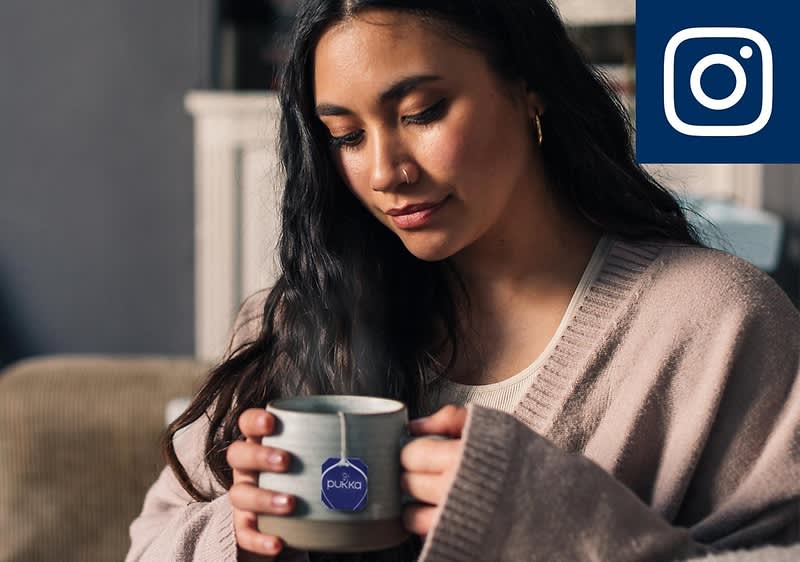 Eine Person in einem beigen Pullover hält eine graue Tasse mit einem Teebeutel der Marke Pukka