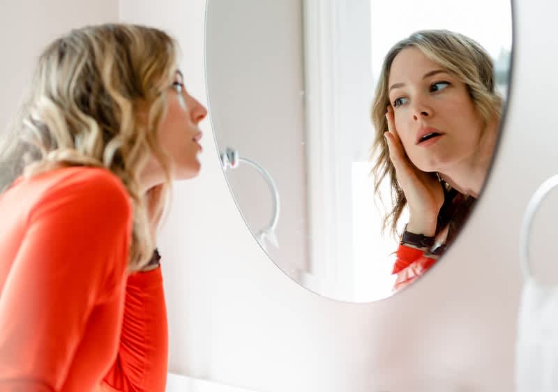 Eine Person mit langen, gewellten Haaren betrachtet sich in einem runden Spiegel. Sie trägt ein rotes Oberteil.