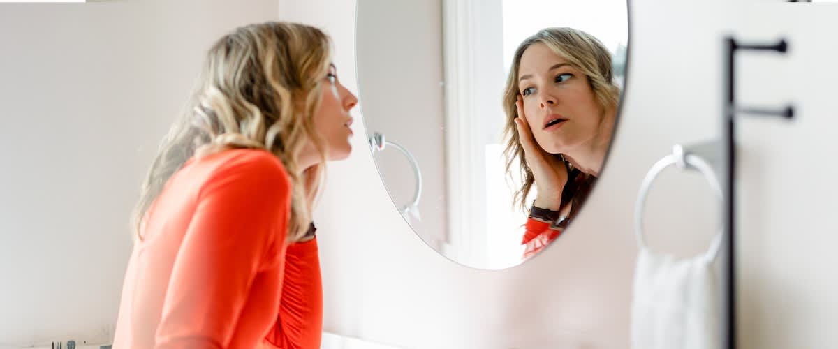 Eine Person mit langen, gewellten Haaren betrachtet sich in einem runden Spiegel. Sie trägt ein rotes Oberteil.