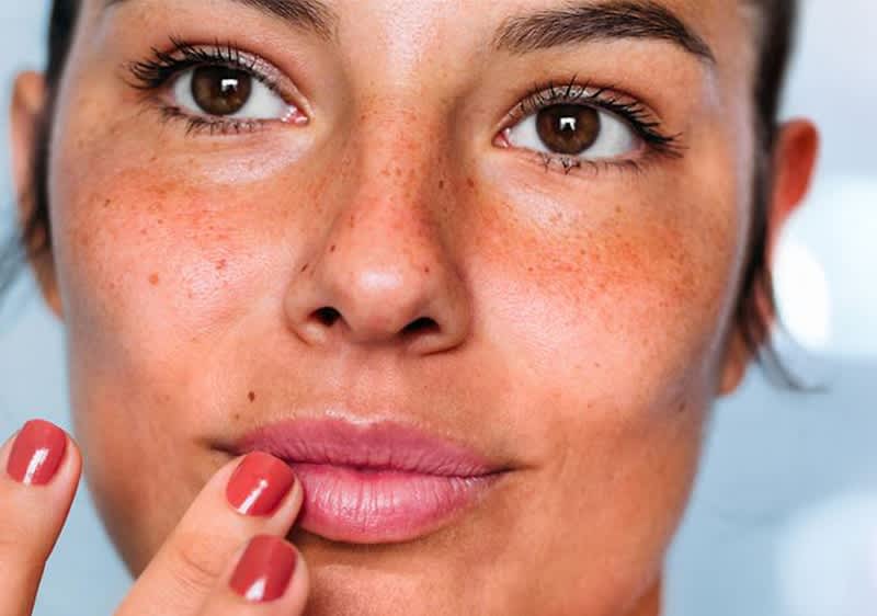 Eine Person hält einen roten Nagellack in der Hand fasst sich an die Lippen