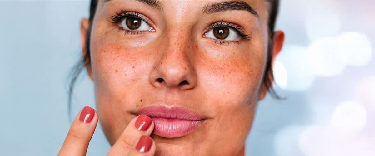 Eine Person hält einen roten Nagellack in der Hand fasst sich an die Lippen