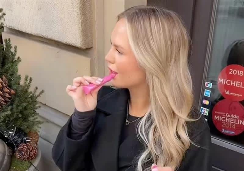 Eine Person mit langen, welligen Haaren hält einen rosa Gegenstand in der Hand. Im Hintergrund sind ein Türrahmen und ein rotes Schild mit der Aufschrift „Michelin Guide 2014“ zu sehen. Links steht ein kleiner Baum mit Zapfen.