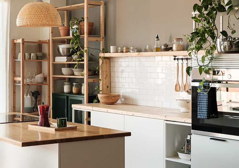 Eine moderne Küche mit Holzregalen, weißen Schränken und einer Arbeitsplatte aus Holz. Pflanzen hängen von der Decke, und auf den Regalen stehen Geschirr und Vorratsgläser. Ein Ofen ist in die Schränke integriert.