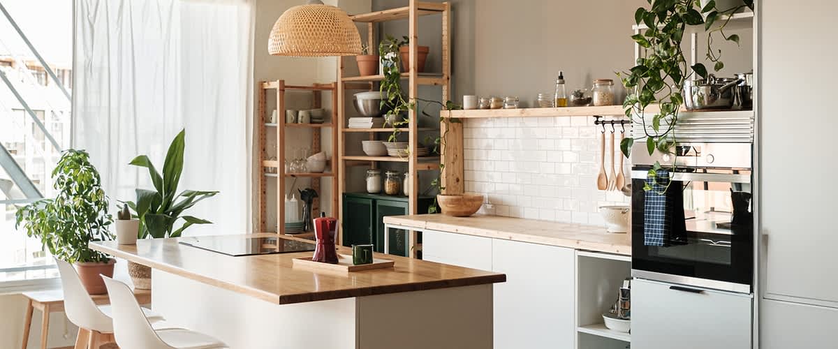 Eine moderne Küche mit Holzregalen, weißen Schränken und einer Arbeitsplatte aus Holz. Pflanzen hängen von der Decke, und auf den Regalen stehen Geschirr und Vorratsgläser. Ein Ofen ist in die Schränke integriert.