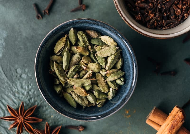 Eine Schale mit grünen Kardamomkapseln auf einem Tisch, umgeben von Sternanis, Zimtstangen und Nelken
