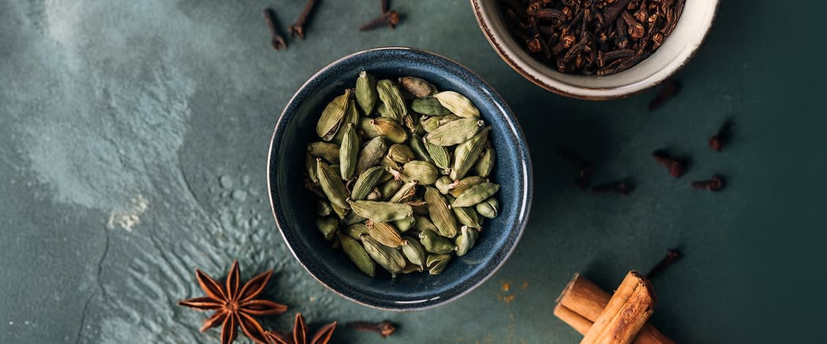 Eine Schale mit grünen Kardamomkapseln auf einem Tisch, umgeben von Sternanis, Zimtstangen und Nelken