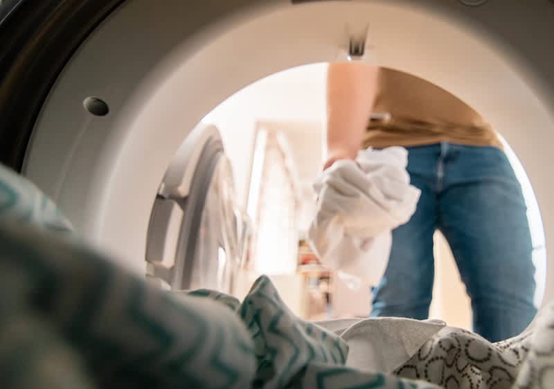 Ein Mann legt Wäsche in eine Waschmaschine, fotografiert aus der Perspektive des Maschineninneren. Im Vordergrund sind bunte Stoffe zu sehen.