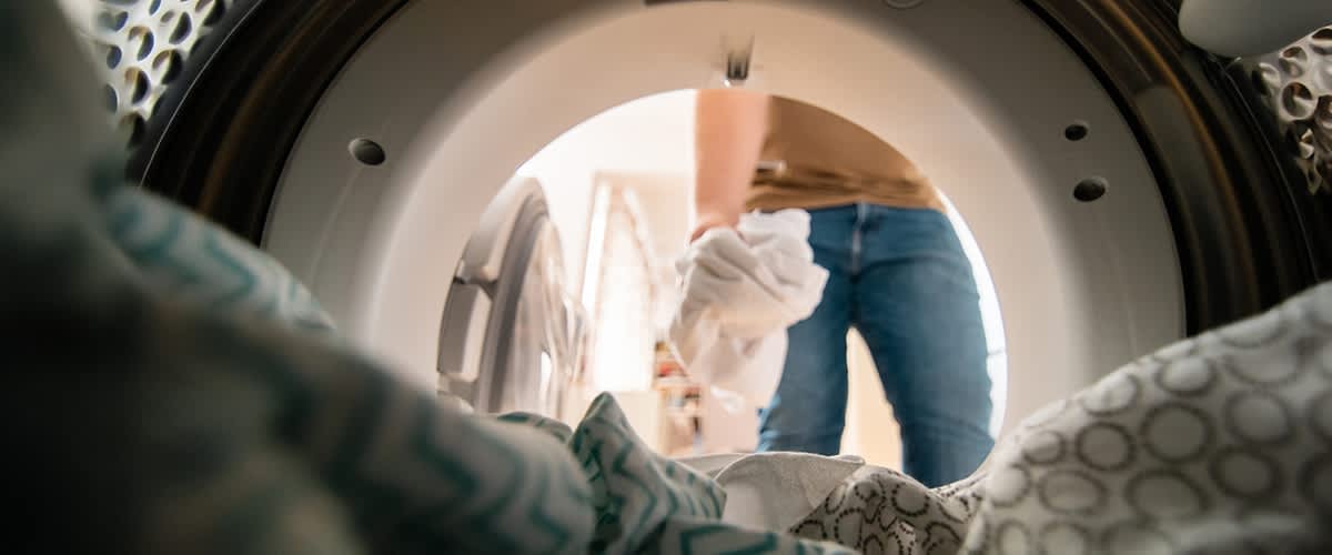 Ein Mann legt Wäsche in eine Waschmaschine, fotografiert aus der Perspektive des Maschineninneren. Im Vordergrund sind bunte Stoffe zu sehen.