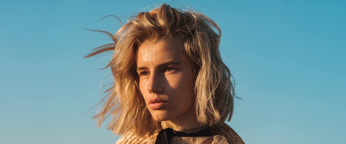 Eine Person mit welligem, blondem Haar steht vor einem blauen Himmel.