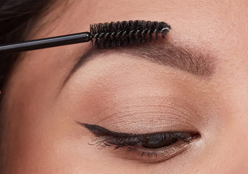 Eine Person verwendet eine Bürste, um ihre Augenbrauen zu kämmen. Das Auge ist mit einem schwarzen Eyeliner betont.