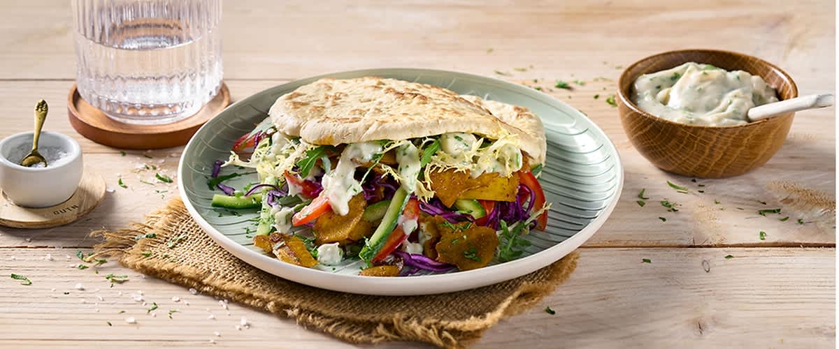 Ein Teller mit einem gefüllten Fladenbrot, daneben eine Holzschale mit cremiger Sauce und ein kleiner Mörser mit Stößel auf einem Holztisch