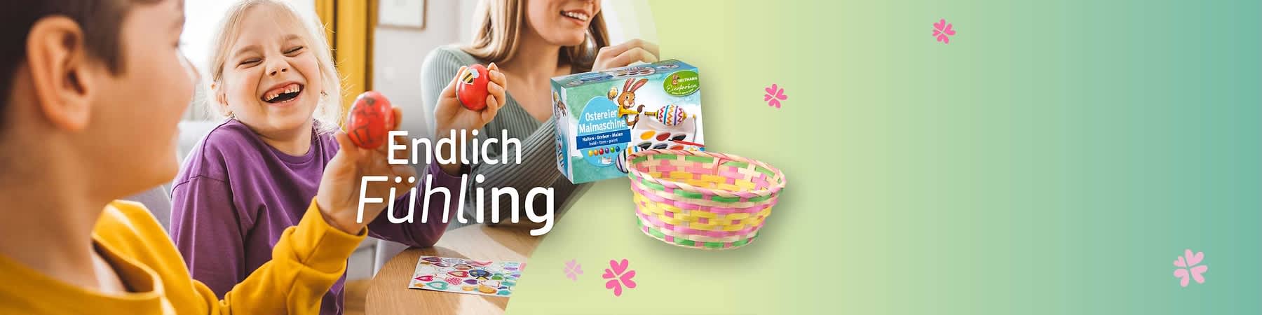 Eine Verpackung mit der Aufschrift „Ostereier Malmaschine“ neben einem bunten Korb aus geflochtenem Material. Im Hintergrund steht der Text „Endlich Frühling“ auf einem grünen Hintergrund mit rosa Blumenmotiven.