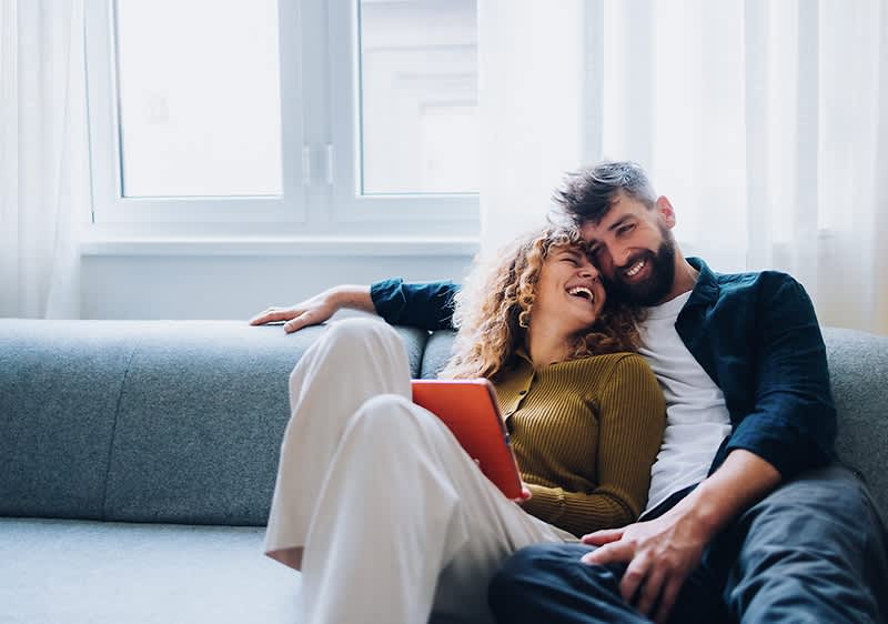 Ein Paar sitzt entspannt auf einem grauen Sofa, die Frau hält ein orangefarbenes Tablet in der Hand.