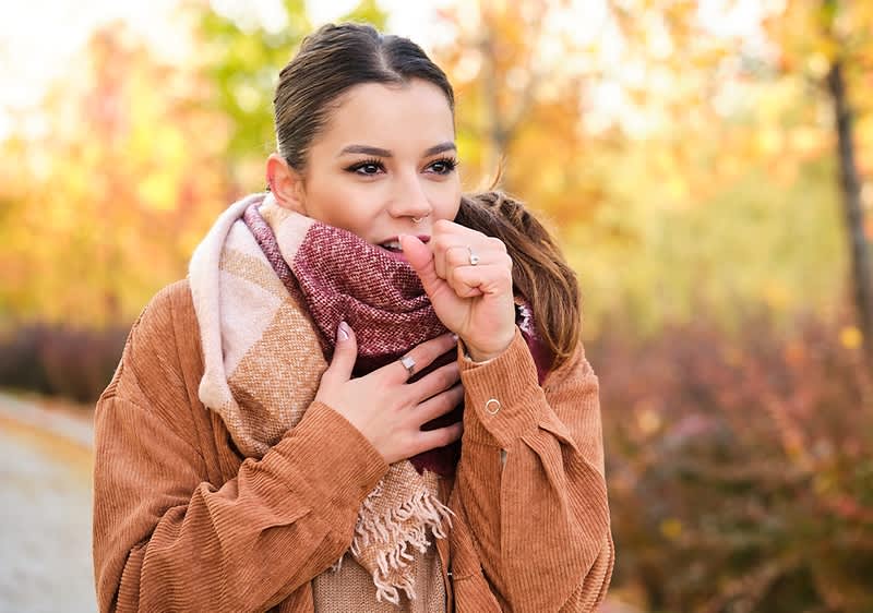 Eine Person in einem braunen Mantel und Schal steht im Freien vor einem herbstlichen Hintergrund mit bunten Bäumen.