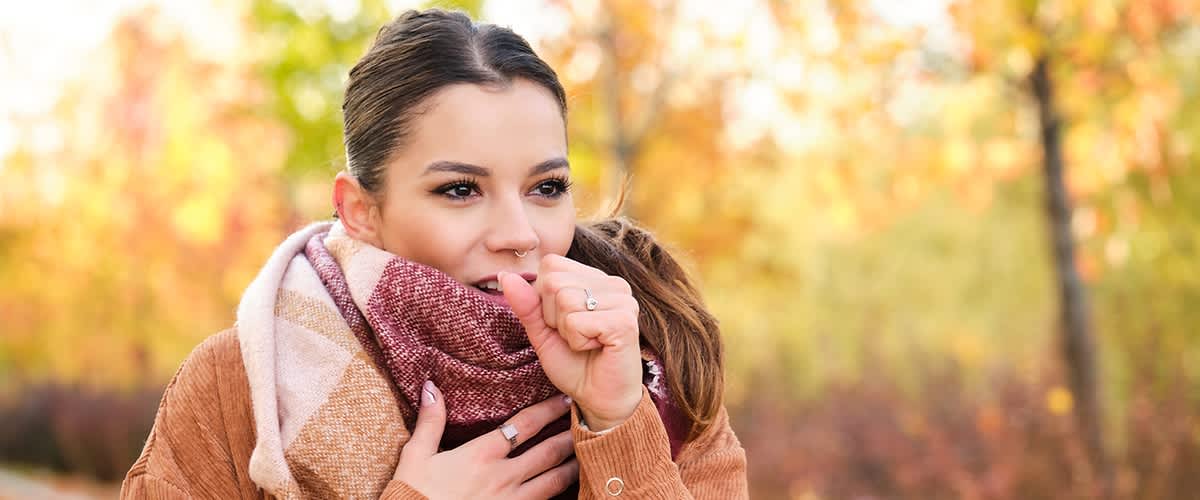 Eine Person in einem braunen Mantel und Schal steht im Freien vor einem herbstlichen Hintergrund mit bunten Bäumen.