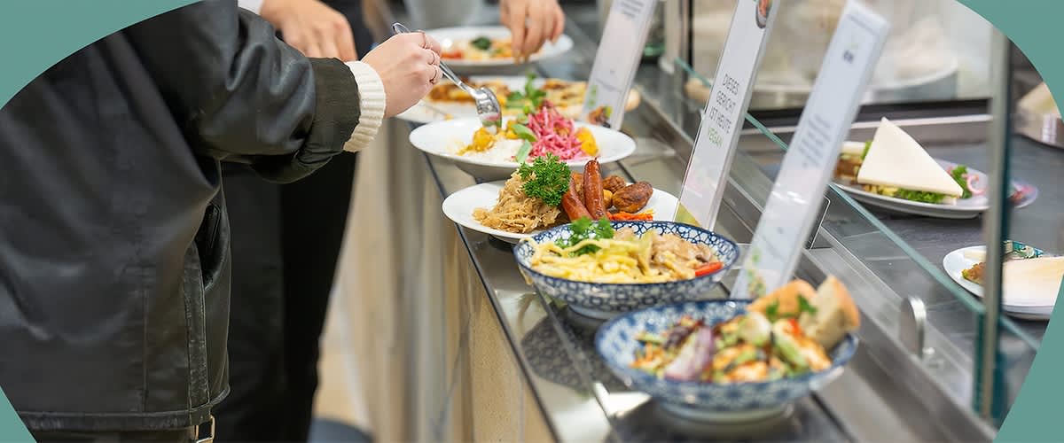Mehrere Teller mit verschiedenen Speisen stehen auf einer Theke, darunter Nudeln, Fleisch und Salat. Eine Person nimmt mit einer Gabel Essen von einem Teller.