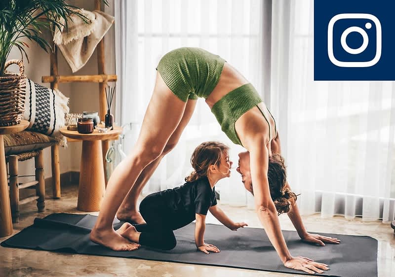 Eine Frau in grüner Sportkleidung macht Yoga in der Position des herabschauenden Hundes, während ein kleines Kind in schwarzer Kleidung unter ihr steht. Im Hintergrund sind ein Holztisch und Pflanzen zu sehen. Oben rechts das Instagram-Logo.