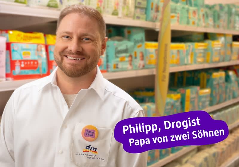 Ein Mann in einem weißen Hemd mit dm-Logo steht vor einem Regal mit Babywindeln. Ein lila Textfeld enthält die Aufschrift „Philipp, Drogist, Papa von zwei Söhnen“