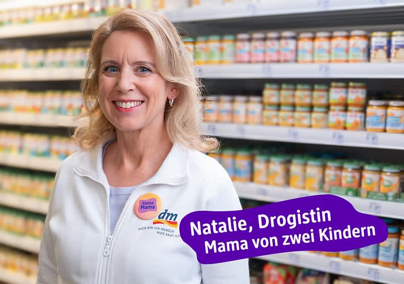 Eine Frau in weißer Arbeitskleidung steht vor einem Regal mit Babynahrung. Auf ihrer Kleidung ist das dm-Logo zu sehen. Daneben steht ein lila Textfeld mit der Aufschrift „Natalie, Drogistin Mama von zwei Kindern“.