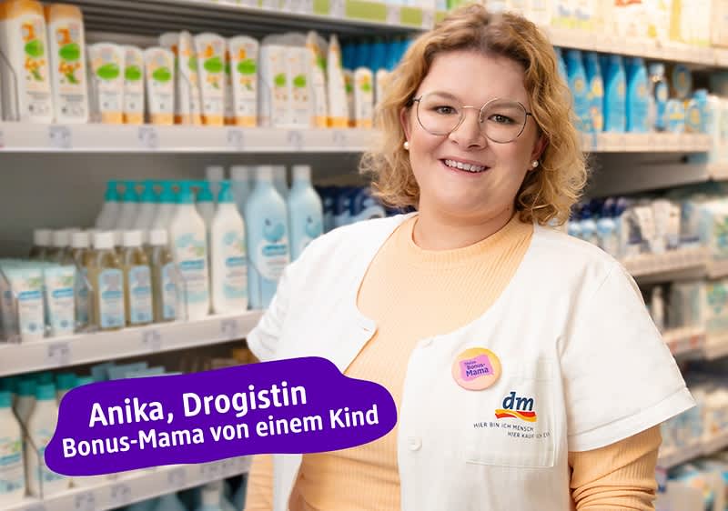 Eine Frau in einem Drogeriemarkt trägt eine weiße Arbeitsjacke mit dem dm-Logo. Im Hintergrund sind Regale mit verschiedenen Pflegeprodukten zu sehen. Ein lila Textfeld enthält die Aufschrift: „Anika, Drogistin, Bonus-Mama von einem Kind“