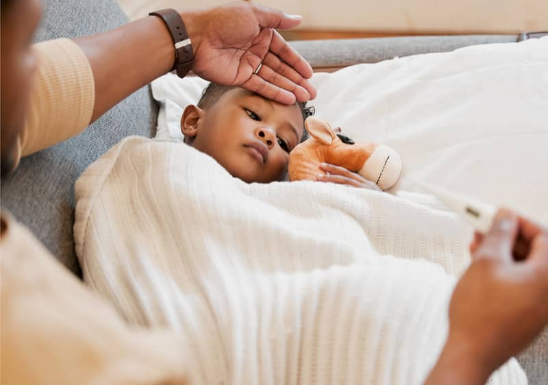 Ein Kind liegt unter einer weißen Decke auf einem Sofa und hält ein Stofftier. Eine Person hält ein Thermometer in der Hand.
