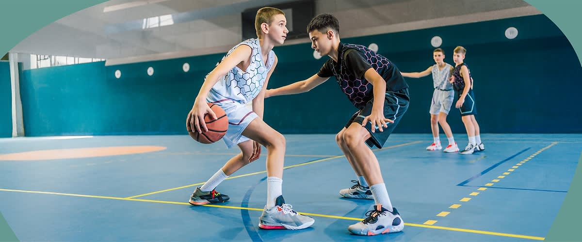 Zwei Jugendliche spielen Basketball in einer Halle, während zwei weitere Jugendliche im Hintergrund stehen und zuschauen.