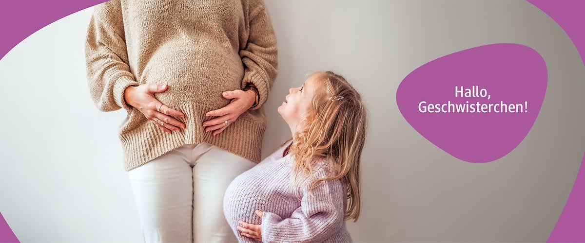 Eine schwangere Frau in einem beigen Pullover steht neben einem kleinen Mädchen, das einen lila Pullover trägt und einen künstlichen Babybauch hat. 