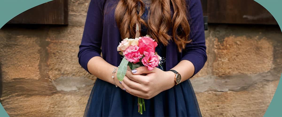 Eine Person mit langen Haaren hält einen kleinen Blumenstrauß mit rosa und weißen Blumen in den Händen, trägt ein dunkelblaues Kleid und eine Uhr am Handgelenk.