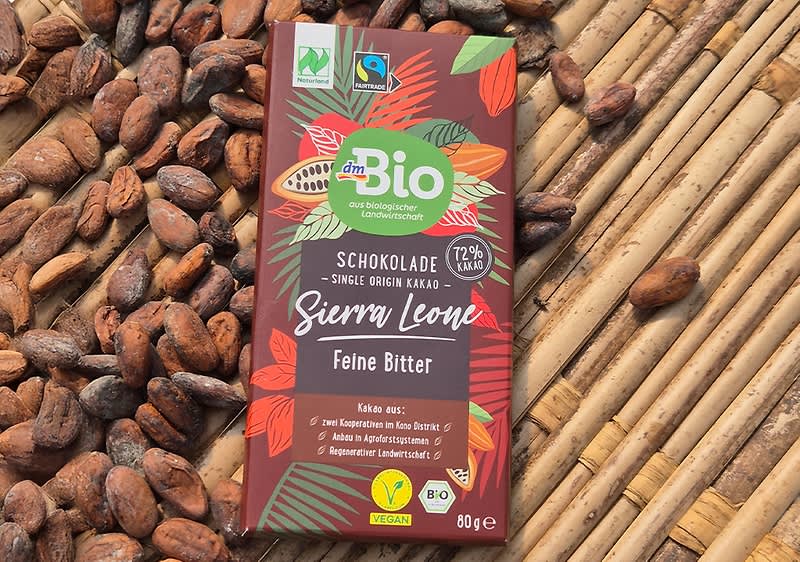 Eine braune Verpackung der dmBio Schokolade „Sierra Leone Fein Bitter“ mit 72 % Kakaoanteil liegt auf Bambusmatten. Die Verpackung zeigt bunte Kakaobohnen und Blätter sowie verschiedene Siegel, darunter Fairtrade und Vegan.