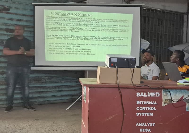 Ein Mann steht neben einer Leinwand, auf der Informationen über die SALMED Cooperative angezeigt werden. Vor der Leinwand sitzt eine Gruppe von Personen an einem Tisch, auf dem ein Projektor und eine Box stehen. Der Tisch ist mit der Aufschrift „SALMED Internal Control System Analyst Desk“ versehen.