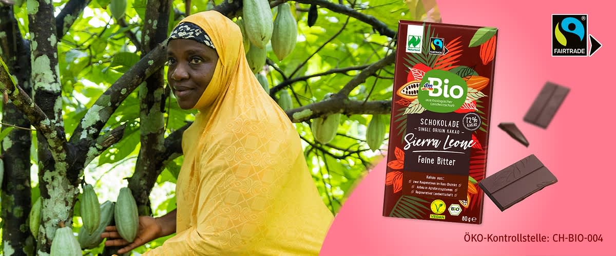 Eine Packung dmBio Schokolade „Sierra Leone Feine Bitter“ mit Fairtrade-Siegel, umgeben von Kakaobohnen und zwei Schokoladenstücken. Text „ÖKO-Kontrollstelle: CH-BIO-004“