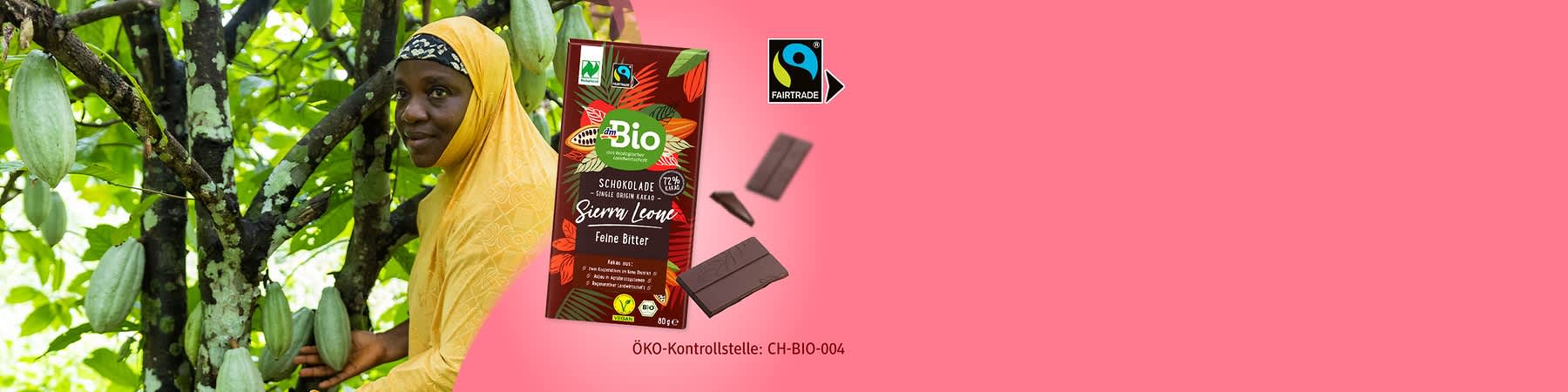 Eine Packung dmBio Schokolade „Sierra Leone Feine Bitter“ mit Fairtrade-Siegel, umgeben von Kakaobohnen und zwei Schokoladenstücken. Text „ÖKO-Kontrollstelle: CH-BIO-004“