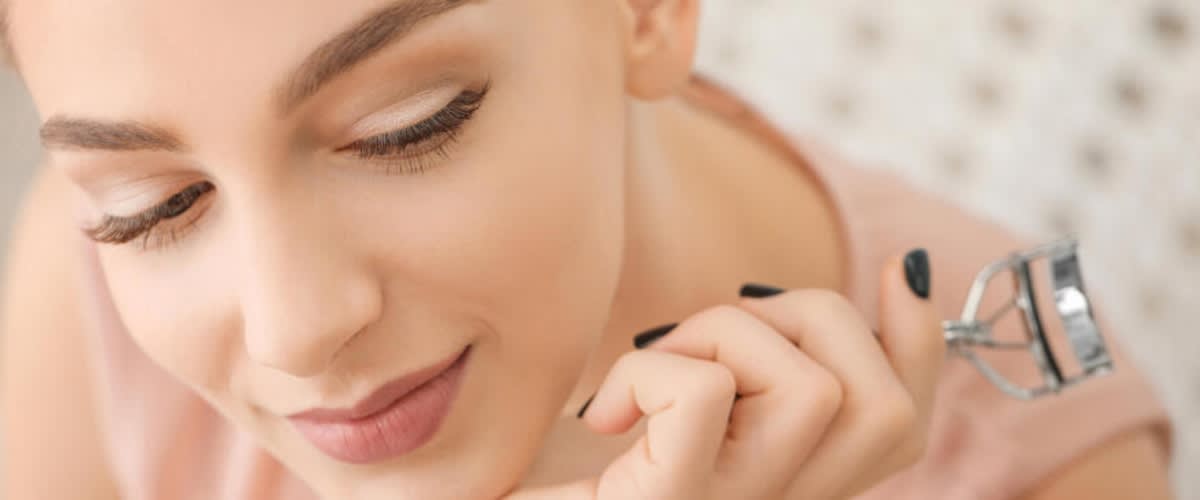 Eine Person hält einen Wimpernzange in der Hand und trägt ein Augen Make-up in dem die Wimpern im Vordergrund stehen