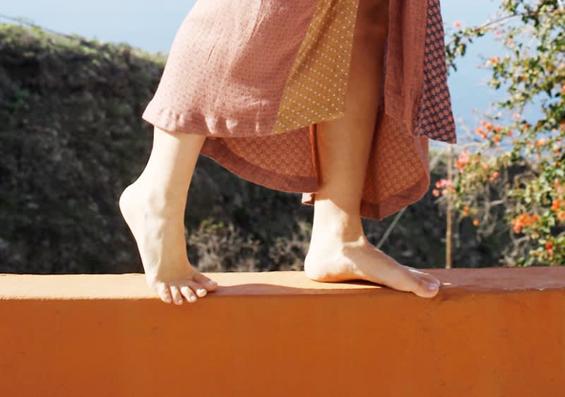 Eine Person geht mit nackten Füßen auf einer orangefarbenen Mauer, trägt dabei ein langes, mehrfarbiges Kleid, im Hintergrund sind Pflanzen zu sehen.