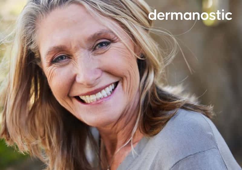 Eine Frau mittleren Alters mit langen Haaren trägt ein graues Oberteil und lächelt. Oben rechts ist das Logo von dermanostic.