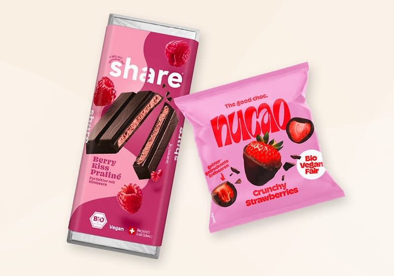 Eine rosa Verpackung von share mit der Aufschrift Berry Kiss Praliné und Bildern von Himbeeren. Daneben eine rosa Verpackung von nucao mit der Aufschrift Crunchy Strawberries und Bildern von Erdbeeren sowie einem Bio- und Vegan-Siegel.