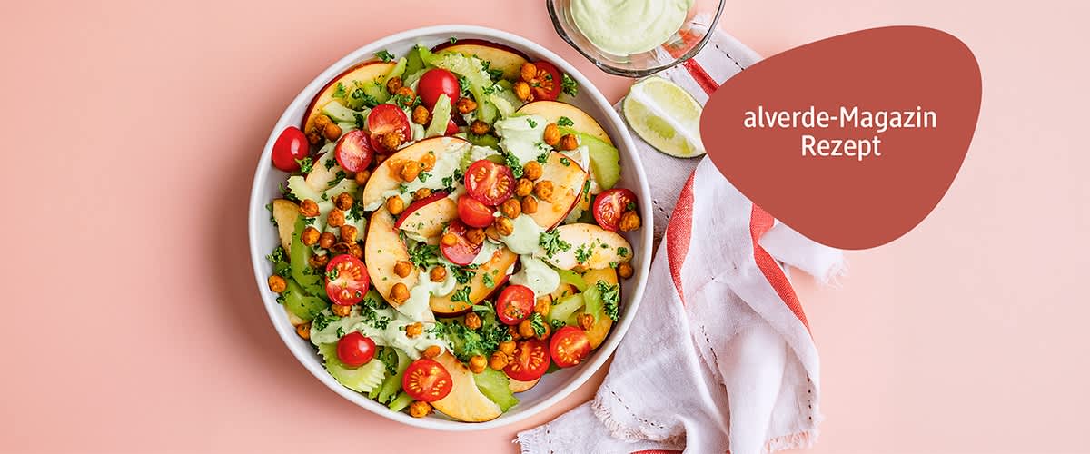 Schüssel mit Salat aus Pfirsichscheiben, halbierten Kirschtomaten, Selleriescheiben, gebackenen Kichererbsen und grüner Kräutercreme, daneben ein Glas mit Dressing und ein weiß-rotes Tuch, oben rechts roter Badge mit Text „alverde-Magazin Rezept“